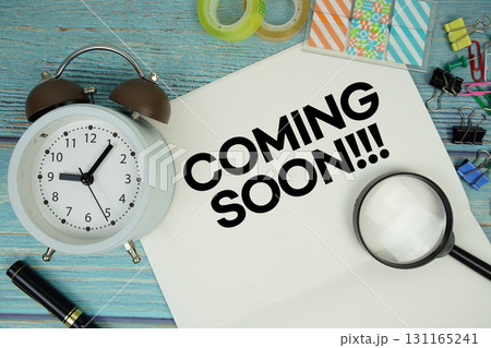 Coming Soon text on a paper surrounded by office supplies, a magnifying glass, and a clock on a blue wooden surface 131165241