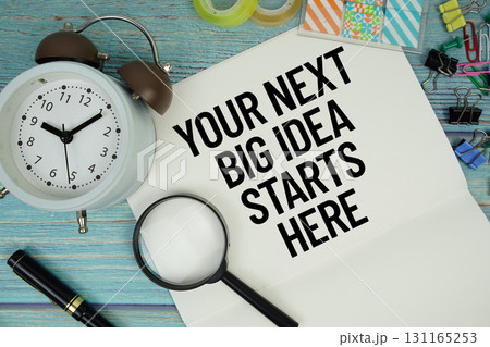 Your Next Big Idea Starts Here text on a paper surrounded by office supplies, a magnifying glass, and a clock on a blue wooden surface Your Next Big Idea Starts Here text on a paper surrounded by office supplies, a magnifying glass, and a clock on a blue wooden surface 131165253