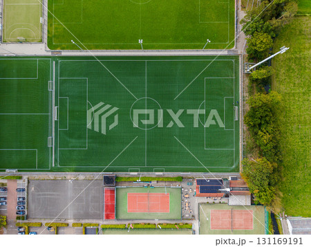 Aerial view of a sports complex with a full size soccer field, smaller fields, red surfaced tennis courts, surrounding buildings, parking areas, and tree lined boundaries. 131169191