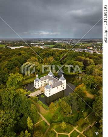 Aerial view of a white turreted castle on a moat island, encircled by dense forest and open fields, with a distant town under a dramatic, cloudy sky. Aerial view of a white turreted castle on a moat island, encircled by dense forest and open fields, with a distant town under a dramatic, cloudy sky. 131169195