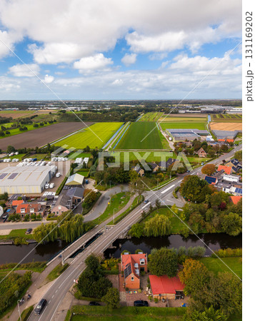 Aerial view of a rural area with a canal and bridge in the foreground, red roofed houses, large warehouse, parked vehicles, and expansive green fields under a partly cloudy sky. Aerial view of a rural area with a canal and bridge in the foreground, red roofed houses, large warehouse, parked vehicles, and expansive green fields under a partly cloudy sky. 131169202