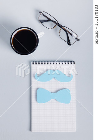 Mustache and bow tie on notebook, coffee mug and glasses on blue top and vertical view 131170183