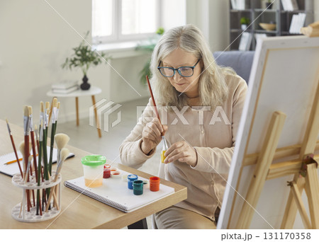Senior woman painting on easel, artist learning to paint on canvas in home living room interior 131170358