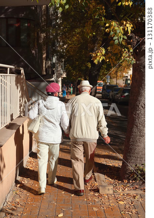 Elderly couple walking hand in hand on autumn street in warm sunlight, dressed in cozy outerwear and hats. Concept of senior love, retirement lifestyle, senior companionship Elderly couple walking hand in hand on autumn street in warm sunlight, dressed in cozy outerwear and hats. Concept of senior love, retirement lifestyle, senior companionship 131170388