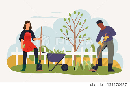 Two gardeners are working in a spring garden, using shovels and a wheelbarrow filled with plants, surrounded by a white picket fence and a newly planted tree 131170427