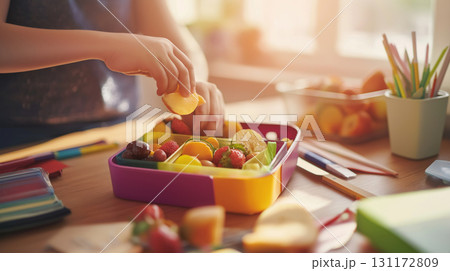 Preparing school lunch on cozy wooden kitchen table with hands placing fruit and snacks into colorful lunchbox, morning light, back to school concept Preparing school lunch on cozy wooden kitchen table with hands placing fruit and snacks into colorful lunchbox, morning light, back to school concept 131172809