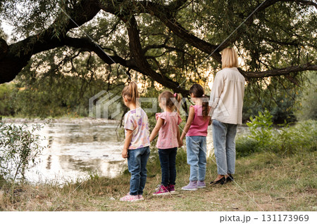 Mother and daughters holding hands looking at river under tree Mother and daughters holding hands looking at river under tree 131173969