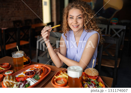 Young woman eating french fries in a restaurant with a table full of food and beer Young woman eating french fries in a restaurant with a table full of food and beer 131174007
