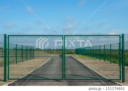Secure metal gate with green fence enclosing private asphalt road leading to open rural landscape 131174602