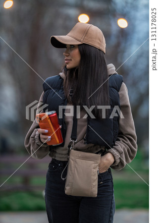 In a serene park setting during autumn a girl with long dark hair and a cap holds her phone. She is dressed in cozy attire enjoying her surroundings as she looks off thoughtfully. In a serene park setting during autumn a girl with long dark hair and a cap holds her phone. She is dressed in cozy attire enjoying her surroundings as she looks off thoughtfully. 131178025