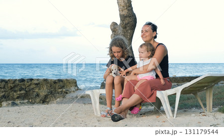 Family gathered on sunbed by ocean cuddling puppy and enjoying happy shared moments  Family gathered on sunbed by ocean cuddling puppy and enjoying happy shared moments  131179544