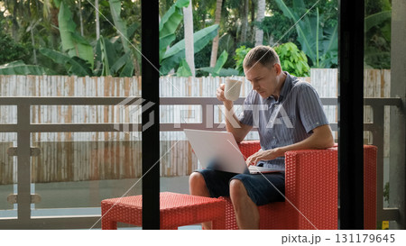 Man enjoys morning coffee while working on a laptop from a balcony surrounded by a lush tropical landscape  131179645