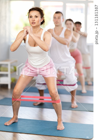 Athletic young woman standing and stretching with resistance bands on mats during workout session with group in gym studio 131181387