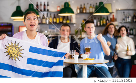 Screaming young adult sports fans rooting for favorite team and waving flag of Uruguay while watching match together in pub Screaming young adult sports fans rooting for favorite team and waving flag of Uruguay while watching match together in pub 131181502