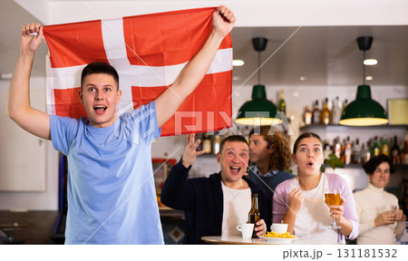 Company of young adult sports fans supporting Danish team with state flag while drinking beer in bar 131181532