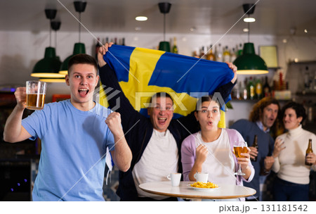 Group of happy friends, sports supporters cheering for favorite team with flag of Sweden while watching match on TV in pub 131181542
