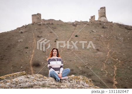 A woman sits on a rock in a field, looking out over the mountains. The sky is cloudy and the landscape is rugged and rocky. The woman is enjoying the view. A woman sits on a rock in a field, looking out over the mountains. The sky is cloudy and the landscape is rugged and rocky. The woman is enjoying the view. 131182047