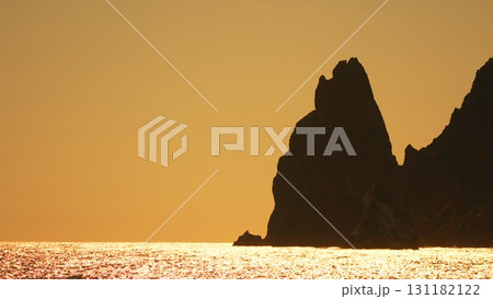 Sea rocks glittering ocean sunset: Coastal cliffs darken as golden sun reflects on sea, creating scenic vista. 131182122