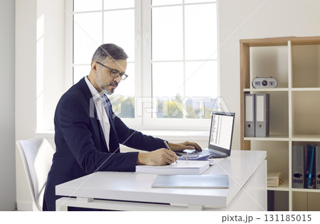 Male accountant or bookkeeper working in office, using laptop, and taking notes in accounting book Male accountant or bookkeeper working in office, using laptop, and taking notes in accounting book 131185015