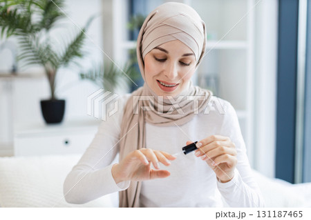 Young Muslim woman wearing head scarf applying nail polish while sitting indoors. Concept of self-care, beauty, and femininity in a modern home environment. Focus on hands and personal grooming. 131187465