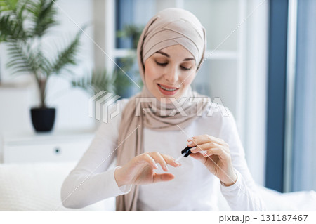 Young Muslim woman wearing head scarf applying nail polish while sitting indoors. Concept of self-care, beauty, and femininity in a modern home environment. Focus on hands and personal grooming. Young Muslim woman wearing head scarf applying nail polish while sitting indoors. Concept of self-care, beauty, and femininity in a modern home environment. Focus on hands and personal grooming. 131187467