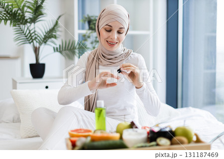 Muslim woman around twenties wearing hijab applies nail oil sitting indoors, surrounded by natural care items such as fruits and oils, portraying a health-conscious lifestyle on self-care and wellness 131187469