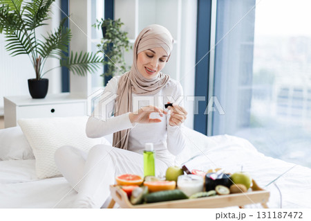 Muslim woman around twenties wearing hijab applies nail oil sitting indoors, surrounded by natural care items such as fruits and oils, portraying a health-conscious lifestyle on self-care and wellness Muslim woman around twenties wearing hijab applies nail oil sitting indoors, surrounded by natural care items such as fruits and oils, portraying a health-conscious lifestyle on self-care and wellness 131187472