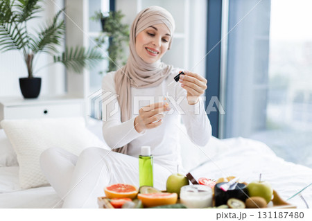Young adult Muslim woman in casual attire using essential oil for healthy nail care. Includes vibrant fruits on table, expressing beauty, wellness, and natural lifestyle within home environment. Young adult Muslim woman in casual attire using essential oil for healthy nail care. Includes vibrant fruits on table, expressing beauty, wellness, and natural lifestyle within home environment. 131187480