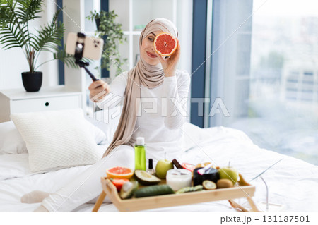 Young Muslim woman wearing hijab filming cosmetic blog using phone in bright room. Tray with skincare products and fruits symbolizes healthy living concept and self-care Young Muslim woman wearing hijab filming cosmetic blog using phone in bright room. Tray with skincare products and fruits symbolizes healthy living concept and self-care 131187501