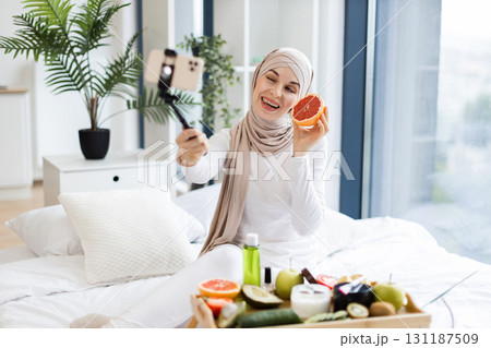 Young Muslim woman in her 20s filming blog with phone about grapefruit use in skincare. She is sitting on bed holding grapefruit, showing organic products, smiling to camera in a light cozy room. Young Muslim woman in her 20s filming blog with phone about grapefruit use in skincare. She is sitting on bed holding grapefruit, showing organic products, smiling to camera in a light cozy room. 131187509