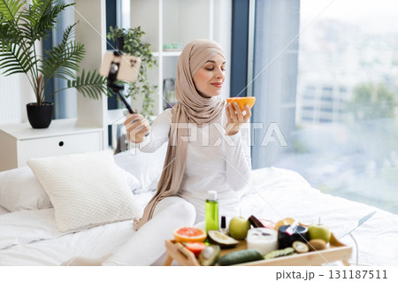 Young Muslim woman wearing hijab filming video blog about skincare using phone indoors. Lady showcasing creative homemade uses of grapefruit for cosmetics while sitting surrounded. Young Muslim woman wearing hijab filming video blog about skincare using phone indoors. Lady showcasing creative homemade uses of grapefruit for cosmetics while sitting surrounded. 131187511