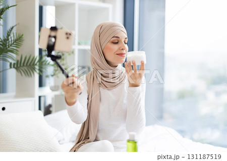 Young Muslim woman wearing hijab demonstrating and reviewing skincare cream with smartphone and tripod indoors in natural light. Focused on facial care routines, modern trends. Young Muslim woman wearing hijab demonstrating and reviewing skincare cream with smartphone and tripod indoors in natural light. Focused on facial care routines, modern trends. 131187519