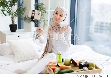 Adult Muslim woman wearing hijab filming content using phone about facial cream and skincare. Tray with fruits and cosmetics visible on bed in modern, bright room. 131187521
