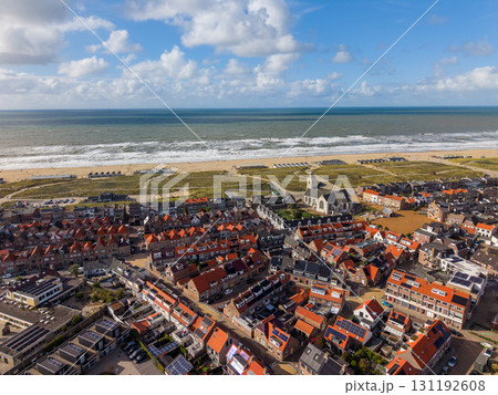 Aerial view of a coastal town with dense red roofed buildings, a tall steepled church, green spaces, wide sandy beach with huts, and ocean waves under a partly cloudy sky. Aerial view of a coastal town with dense red roofed buildings, a tall steepled church, green spaces, wide sandy beach with huts, and ocean waves under a partly cloudy sky. 131192608