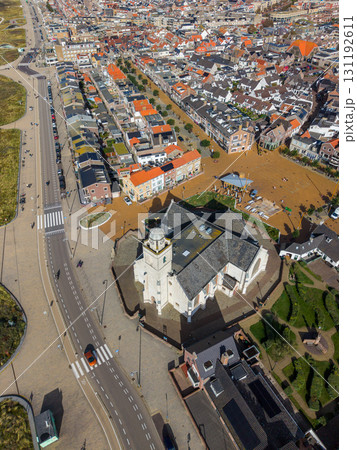 Aerial view of a compact town centered on a white facade church with dome and clock tower, surrounded by cobblestone streets, red and gray roofed buildings, green spaces, and parked cars. Aerial view of a compact town centered on a white facade church with dome and clock tower, surrounded by cobblestone streets, red and gray roofed buildings, green spaces, and parked cars. 131192611