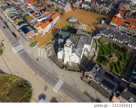 Aerial view of a compact town centered on a white facade church with dome and clock tower, surrounded by cobblestone streets, red and gray roofed buildings, green spaces, and parked cars. Aerial view of a compact town centered on a white facade church with dome and clock tower, surrounded by cobblestone streets, red and gray roofed buildings, green spaces, and parked cars. 131192612