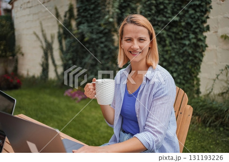 Woman working remotely from her garden, enjoying a cup of coffee Woman working remotely from her garden, enjoying a cup of coffee 131193226