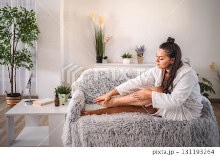 Woman applying moisturizing cream on legs sitting on sofa at home 131193264