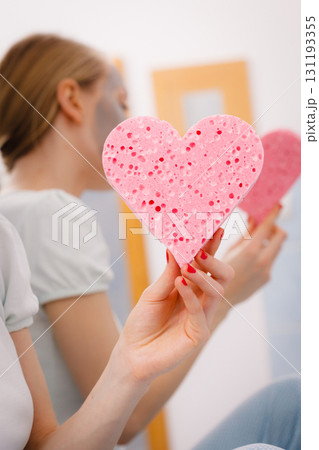 Woman having grey face mask holding heart sponge Woman having grey face mask holding heart sponge 131193355