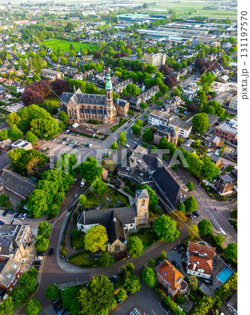 Aerial view of a European town with a tall green spire gothic church at its center, surrounded by red roofed houses, winding streets, trees, and fields blending into rural outskirts. 131197570