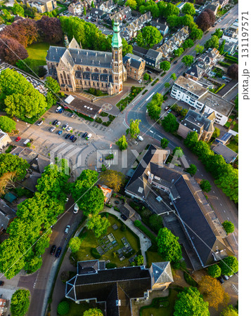 Aerial view of a European town with a tall green spire gothic church at its center, surrounded by red roofed houses, winding streets, trees, and fields blending into rural outskirts. 131197571