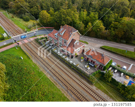 Aerial view of a red roofed cafe beside railway tracks and a road crossing with safety barriers, set in a green semi rural landscape of trees and vegetation. 131197585