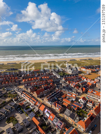 Aerial view of a coastal town with dense red roofed buildings, a tall steepled church, green spaces, wide sandy beach with huts, and ocean waves under a partly cloudy sky. 131197589
