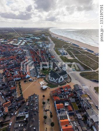 Aerial view of a coastal town with red roofed houses, a white church and bell tower near dunes and beach, main road along the shore, and partly cloudy skies adding depth. Aerial view of a coastal town with red roofed houses, a white church and bell tower near dunes and beach, main road along the shore, and partly cloudy skies adding depth. 131197591