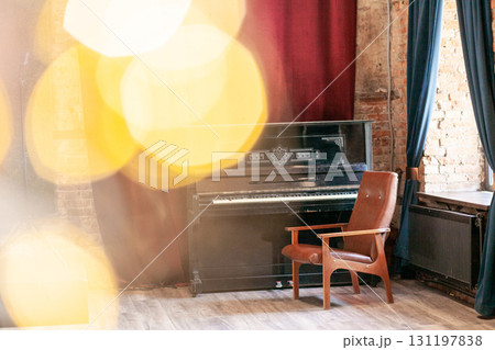 Old vintage piano and armchair stands in the hall against the background of a red curtain and a brick wall near the big window, with bokeh and highlights Old vintage piano and armchair stands in the hall against the background of a red curtain and a brick wall near the big window, with bokeh and highlights 131197838