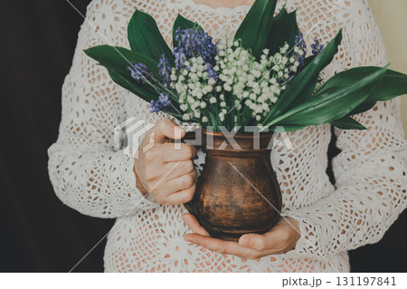 Bouquet of lilies of the valley in ceramic jug in the hands of woman in knitted blouse 131197841