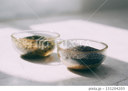 Glass bowls filled with chia seeds and quinoa on white marble surface, sunlight casting soft shadows, healthy food concept, minimal style, natural light 131204203