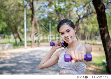 A young woman joyfully exercising outdoors with dumbbells in a sunny park. 131204635