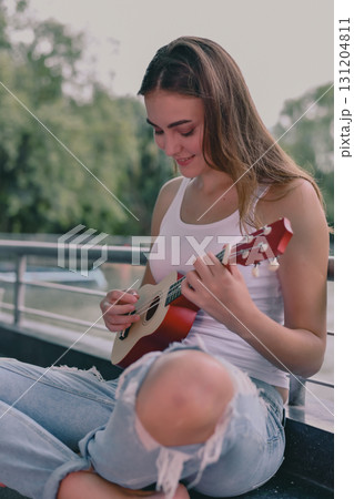 A young woman enjoys playing a ukulele outdoors, expressing joy and creativity in a serene environment. 131204811