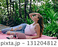 A young woman relaxes outdoors, lying on a blanket, enjoying nature while using a laptop amidst lush greenery. 131204812